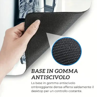 Immagine ravvicinata e descrittiva che mostra la base in gomma antiscivolo di un tappetino da scrivania. Una mano solleva l'angolo per rivelare la superficie inferiore nera e texturizzata. Un cerchio ingrandito evidenzia la fitta trama della gomma, con il testo che recita "BASE IN GOMMA ANTISCIVOLO"