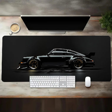Vista dall'alto di un tappetino da scrivania esteso che mostra il profilo laterale di un'auto sportiva classica nera (stile Porsche 930 Turbo) con cerchi oro, su uno sfondo nero. Una tastiera bianca e un mouse sono posizionati sul tappetino, parte di un setup da scrivania con monitor, tazza di caffè e una pianta su una scrivania di legno.